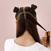 a person with long brown hair uses the dyson airstrait while standing in front of a pink background