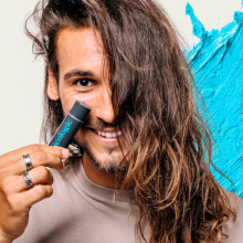 Person holding geologie stick up to face and smiling with swatch of blue product behind them