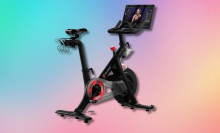 A black Peloton stationary bike with a red wheel and a large touchscreen displaying a fitness instructor.