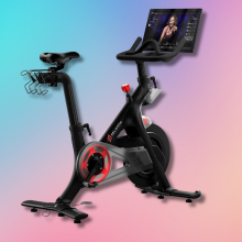 A black Peloton stationary bike with a red wheel and a large touchscreen displaying a fitness instructor.