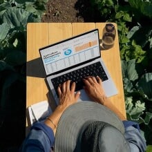 an aerial photo shows a person uses the macbook pro while wearing a bucket hat and sitting in a field of cabbage