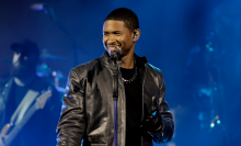 Usher smiles in front of a mic and stand. He wears a black leather jacket and gloves 