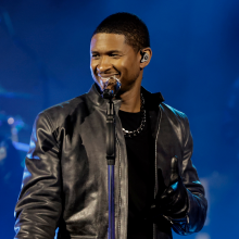 Usher smiles in front of a mic and stand. He wears a black leather jacket and gloves 