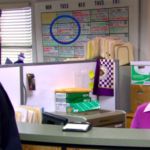 Pam sits at her desk and talks to Michael who is standing at reception.