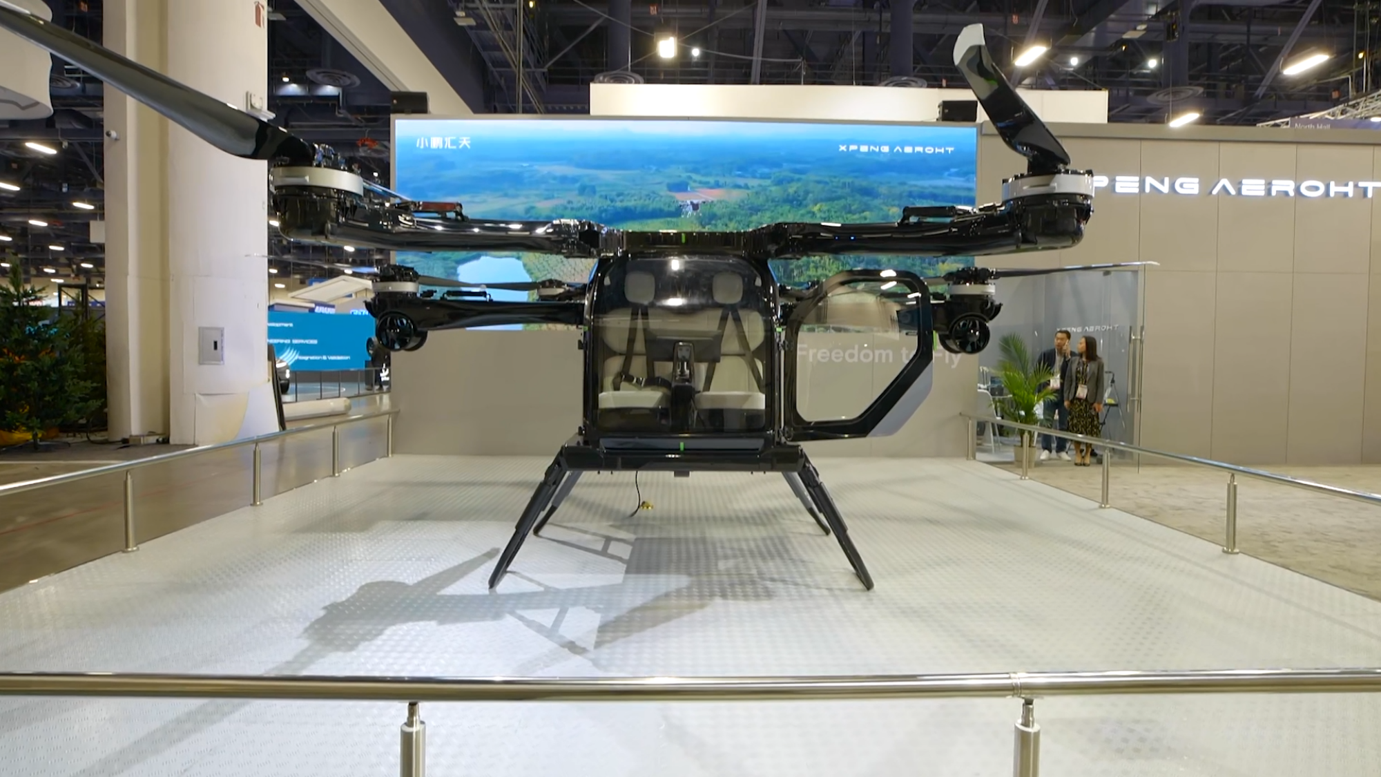 A futuristic flying vehicle displayed in an exhibition setting, featuring a cockpit with two seats, quadcopter-style rotors, and landing gear. 