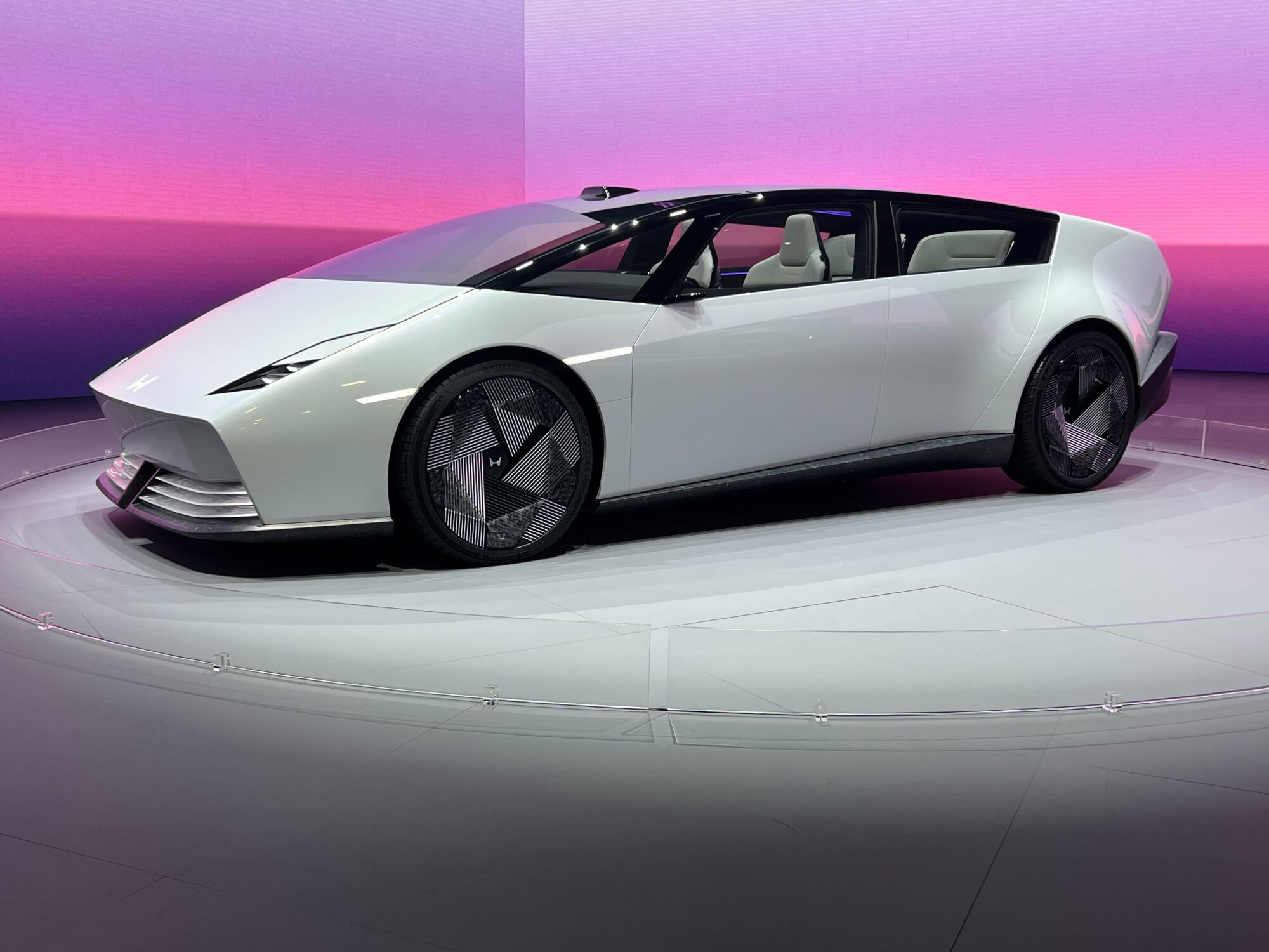 A sleek white concept car with a futuristic design, sharp lines, and unique wheel patterns displayed on a platform against a gradient purple and pink background, suggesting an unveiling or showcase event.