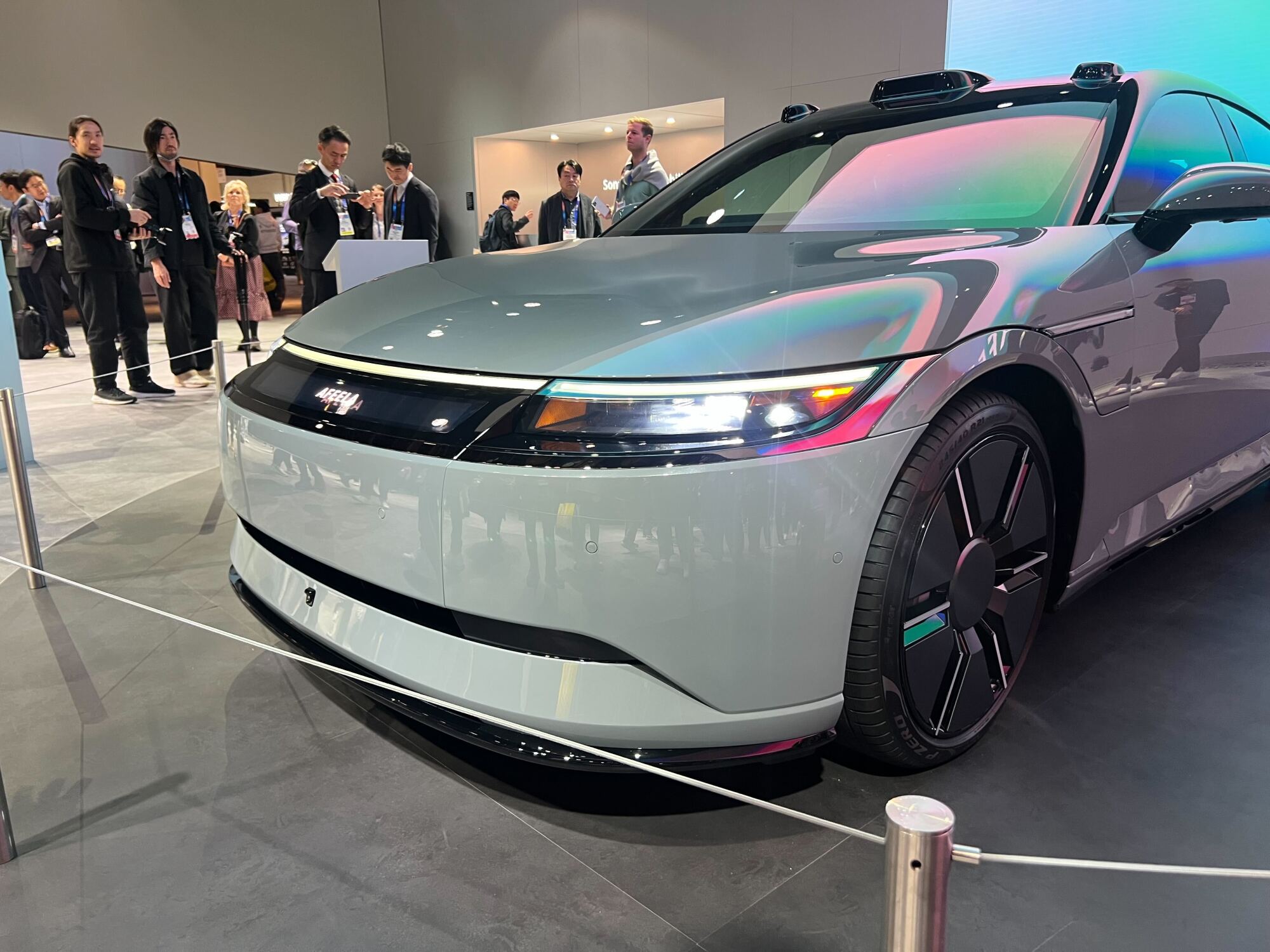 A close-up view of the front of an AFEELA electric car on display, showcasing its sleek aerodynamic design, illuminated headlights, and a high-tech aesthetic. The reflective surface highlights ambient lighting, while a group of attendees observes the vehicle in the background at an exhibition.