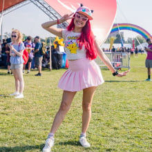 A women in a light pink pleated skirt, white sneakers, and cap. She has red hair and is making a peace sign with her left 