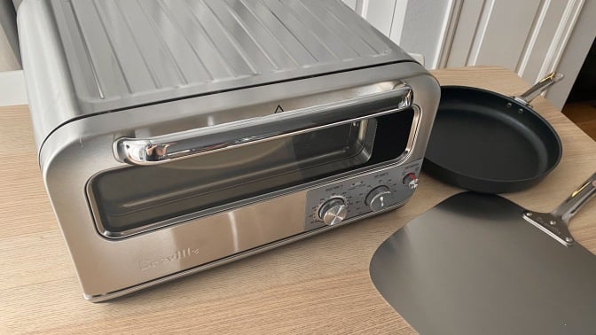 Breville pizza oven on a wood table next to a pizza pan and a peel