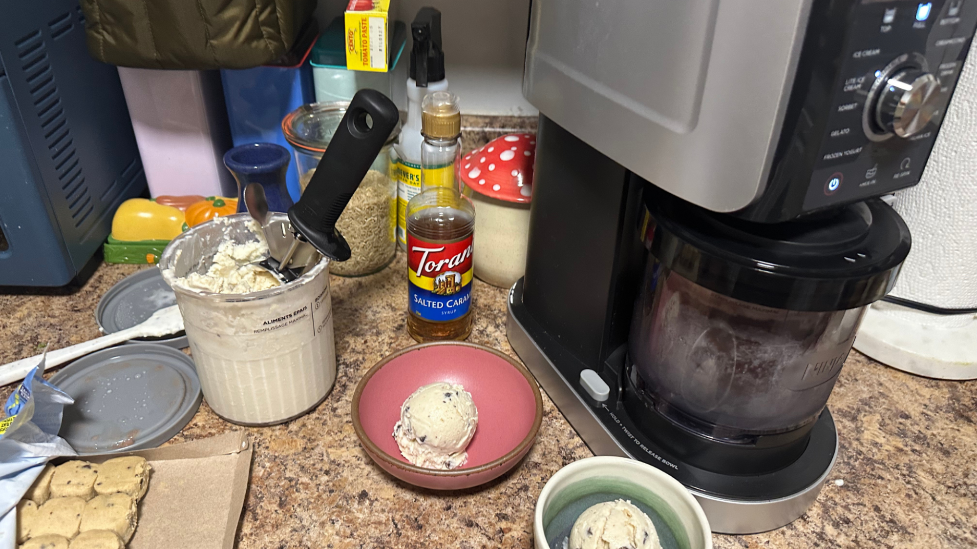 On the counter, the Ninja Creami next to a pint of ice cream and scoops of the ice cream in bowls.