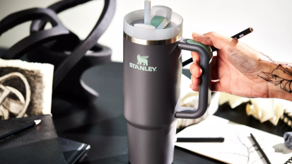 a close-up of a tattooed person holding a black stanley quencher tumbler on a cluttered desk