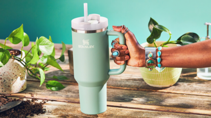 a close-up of a woman wearing turquoise jewelry holding a stanley quencher cup on a wooden table amid potted plants