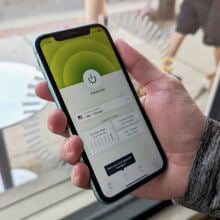 a close-up of a man holding an iphone displaying the expressvpn app in front of a window