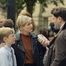 Jodie Whittaker stands in a scene from "Toxic Town" as a mother with her son talking to a reporter.