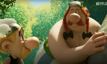 Two animated characters stand in the forest beside a boulder.