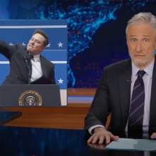 A man sits in front of a talk show desk. In the top-left is an image of another man appearing to give a Nazi salute.