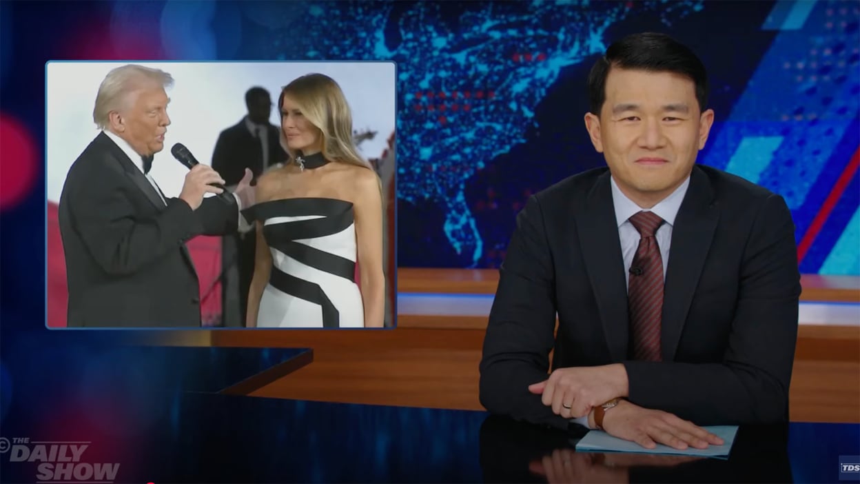 A man in a suit sits behind a talk show desk. In the top-left is an image of a man and woman in party dress.