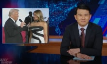 A man in a suit sits behind a talk show desk. In the top-left is an image of a man and woman in party dress.