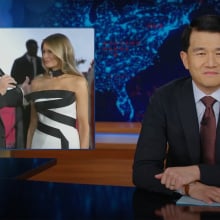 A man in a suit sits behind a talk show desk. In the top-left is an image of a man and woman in party dress.