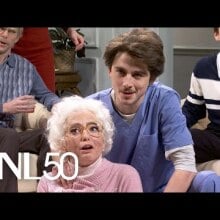 family crowded around a woman in elderly makeup. Next to her is white man in blue nursing scrubs