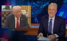 A man sits behind a talk show desk, grinning. In the top-left is an image of another man in a suit.