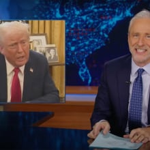 A man sits behind a talk show desk, grinning. In the top-left is an image of another man in a suit.