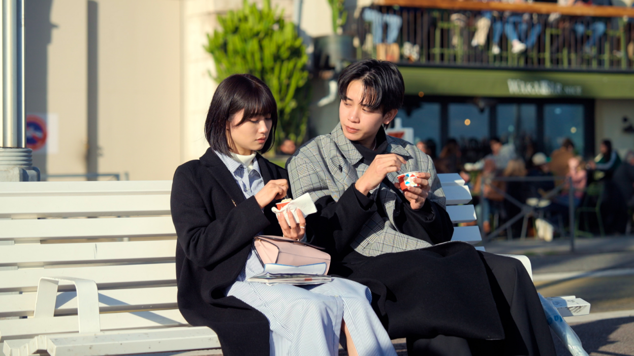 A still from 'Offline Love' showing a man and a woman eating ice cream on a bench. The man is looking at the woman, and the woman is looking at her ice cream.