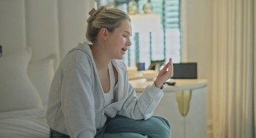 person sitting on a bed in a lightfilled room gesturing with their hand