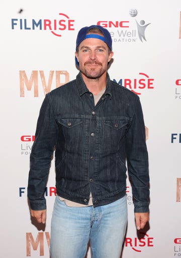 stephen amell stands looking at the camera with a neutral facial expression, he has brown hair and beard, he wears a cap backwards, grey tshirt, blue denim jacket and jeans