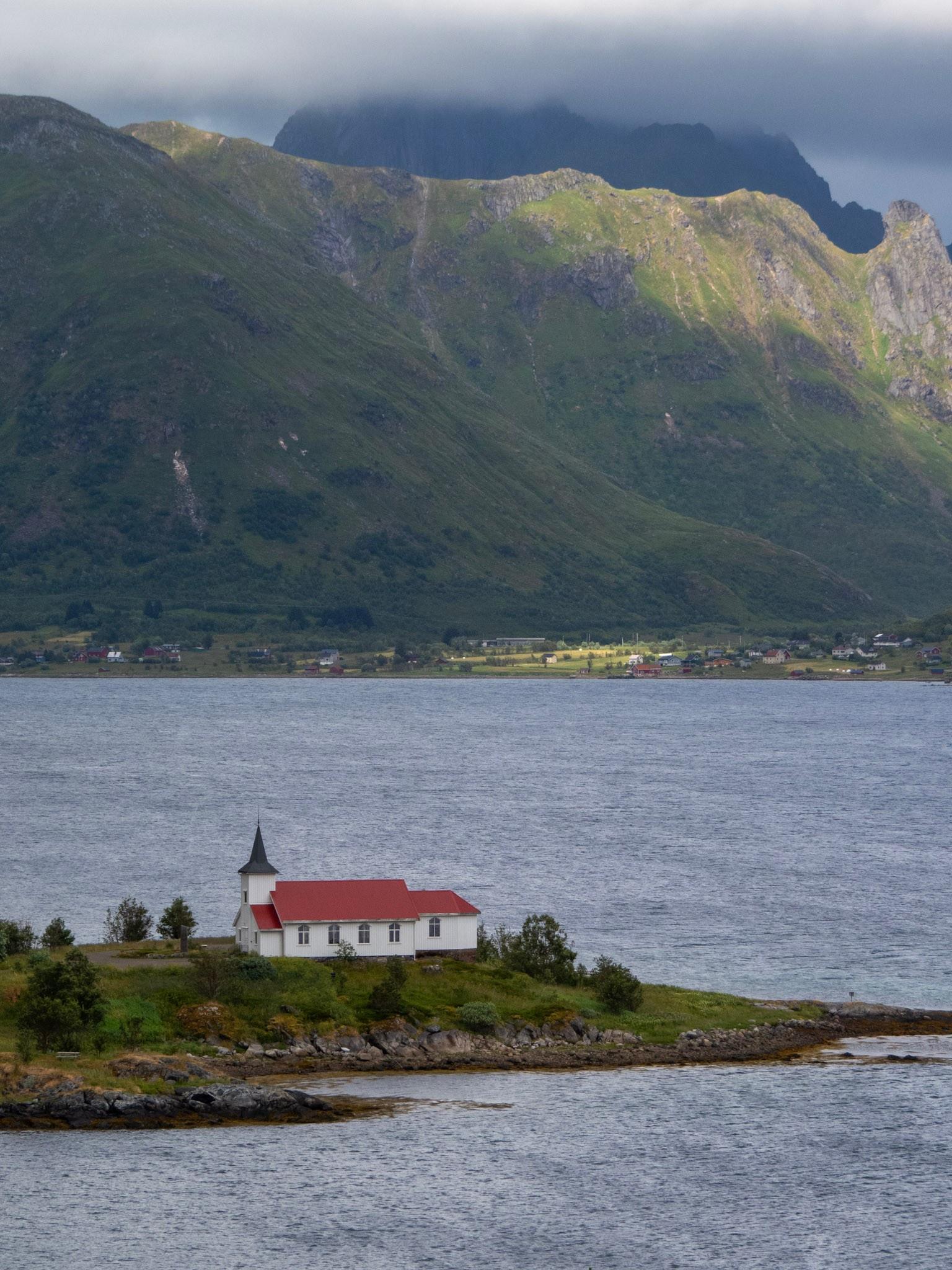 A small white church sits below on a spit of land in the sea. Behind it dramatic peaks spear into clouds, sun pouring through a few gashes.