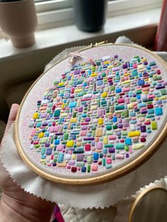 a person holding up a colorful cross stitch pattern on a white piece of cloth in front of a window