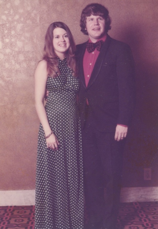 A woman with long brunette hair wearing a long green dress with white polka dots stands to the left of a man with brunette hair wearing a brown suite with a red shirt. Both are smiling broadly.