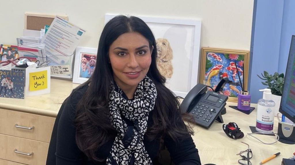 Dr Deepali Misra-Sharp sits at her work desk, with pictures and cards behind her.