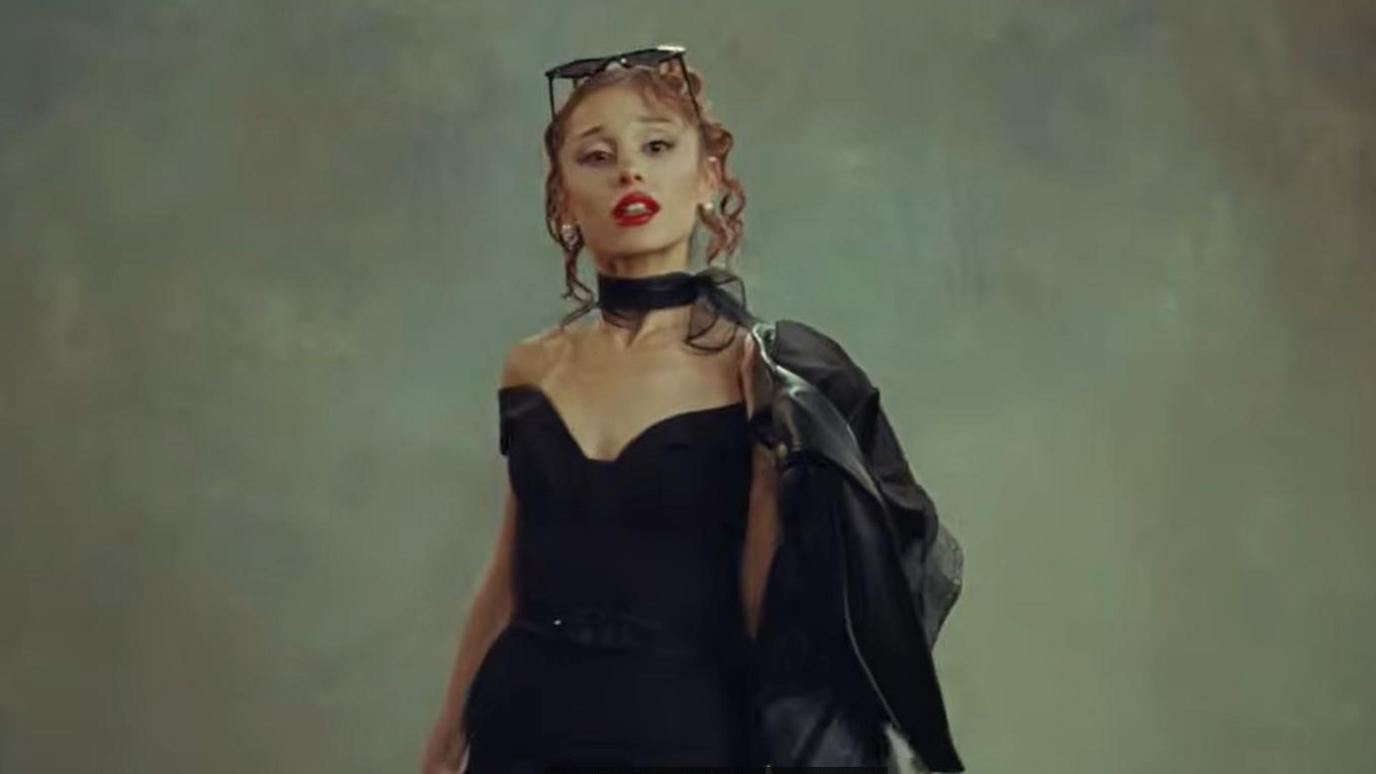 Ariana Grande in a black dress with a black biker jacket over one shoulder and dark glasses on her head.