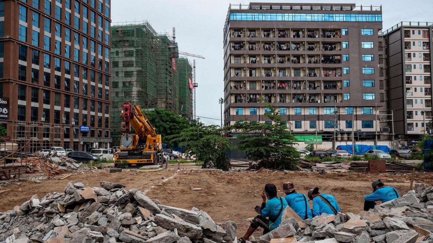 Workers in blue shirts sit amid construction rubble in front of tall buildings near a crane.
