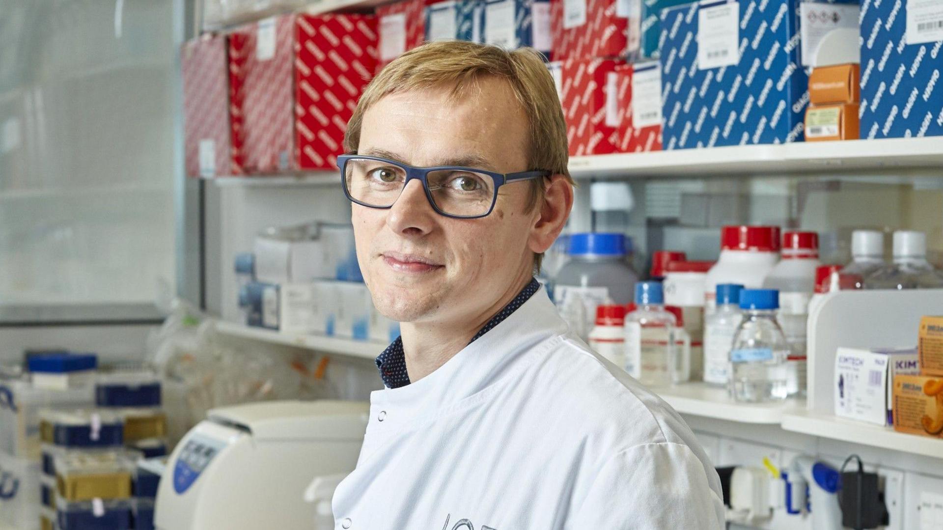 Professor Graham is standing in a laboratory setting in front of shelves full of jars and boxes. He is looking directly at the camera, wearing a white lab coat and glasses, with blondish hair