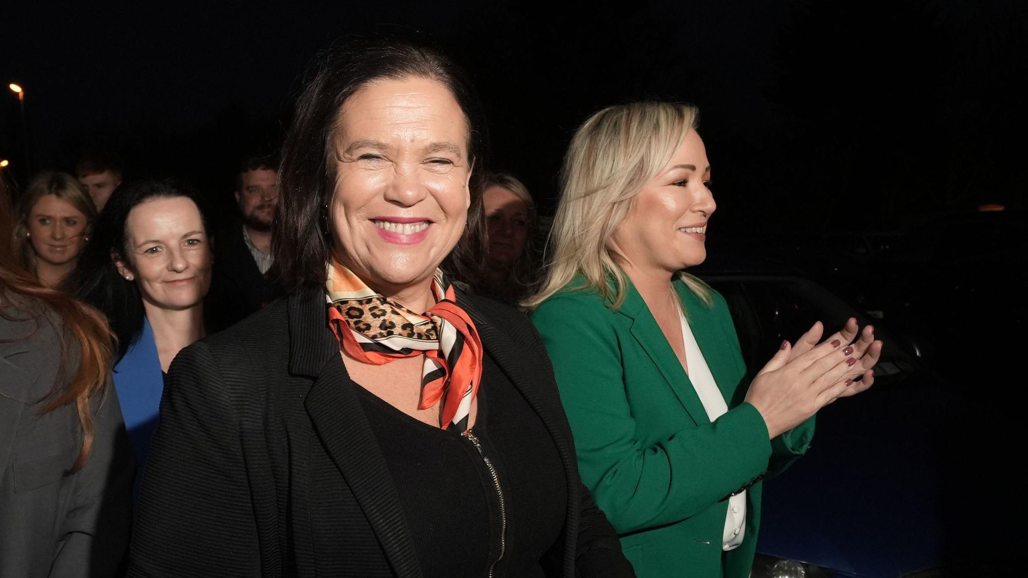 A woman with dark brown chin length hair smiles towards the camera. She is wearing a black top and jacket with a red patterned scarf. A woman with shoulder length blond hair stands to herright wearing a green blazer and white top. She is smiling to the right of the lens and clapping her hands. 