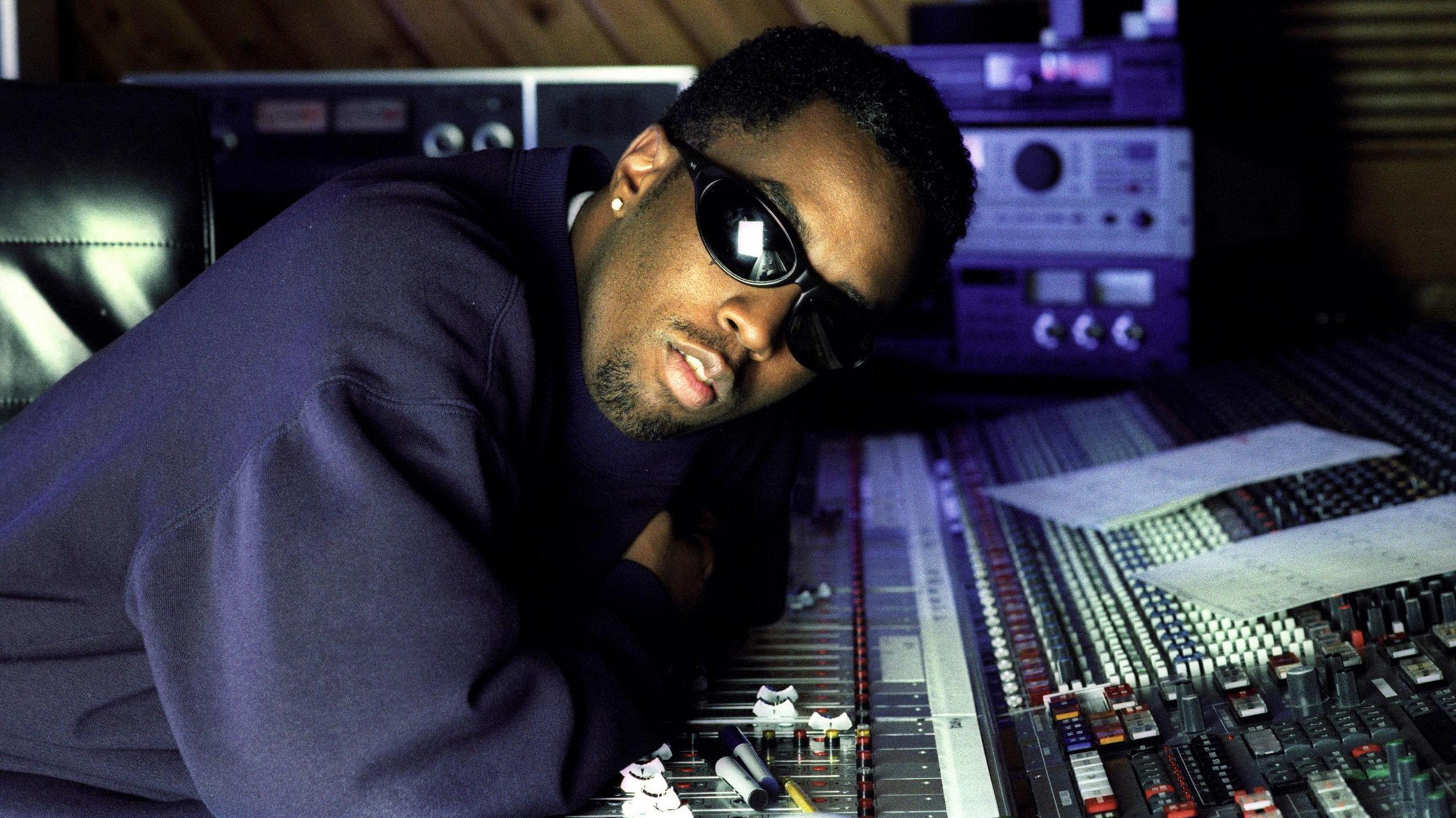 Sean "Diddy" Combs, wearing sunglasses and a baggy navy sweatshirt, with an stud in his right ear, neatly trimmed hair and a goatee, leaning over the mixing desk of his recording studio, which is strewn with papers and Sharpie pens