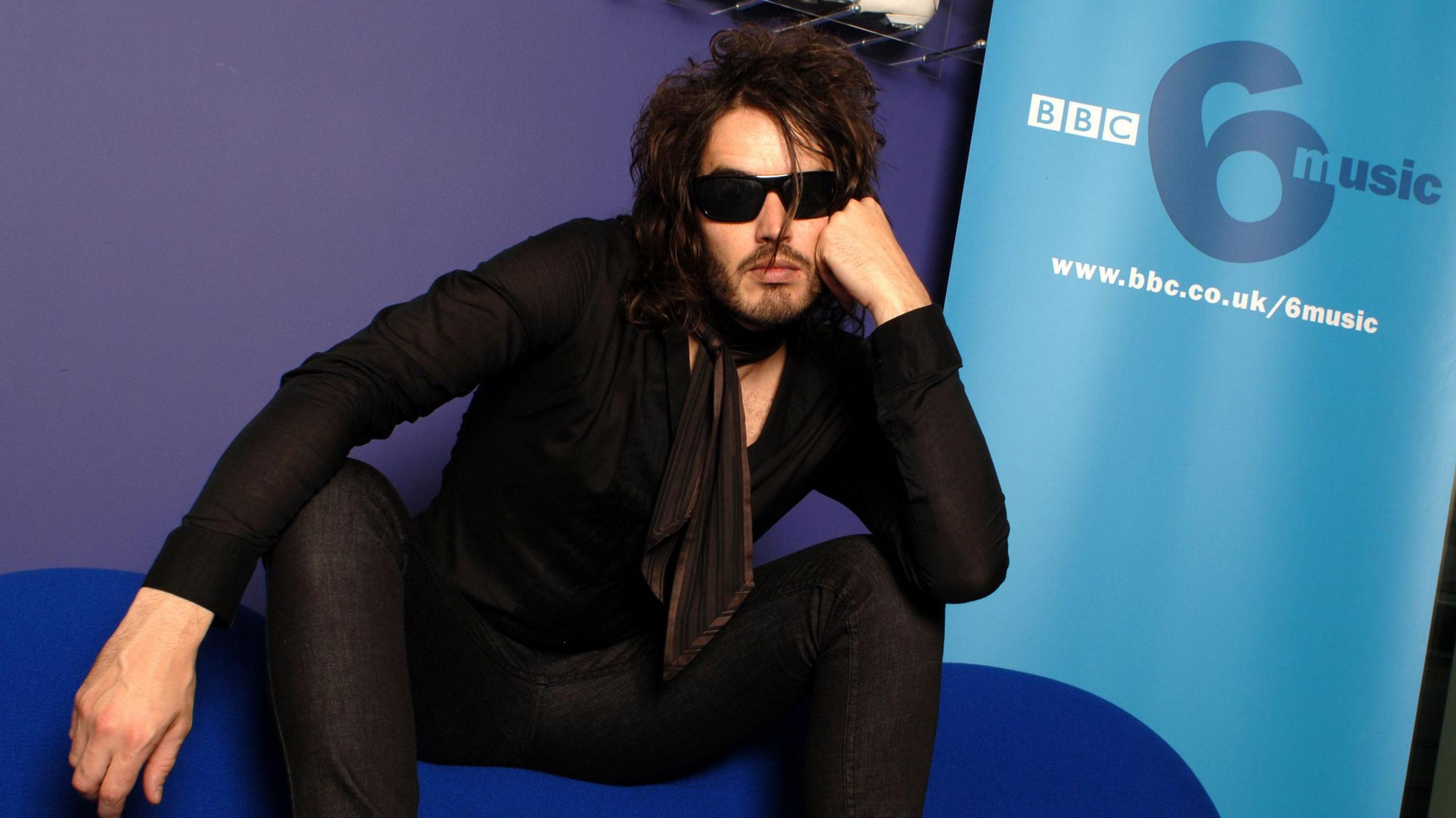 Russell Brand wearing dark glasses in front of 6 Music branding