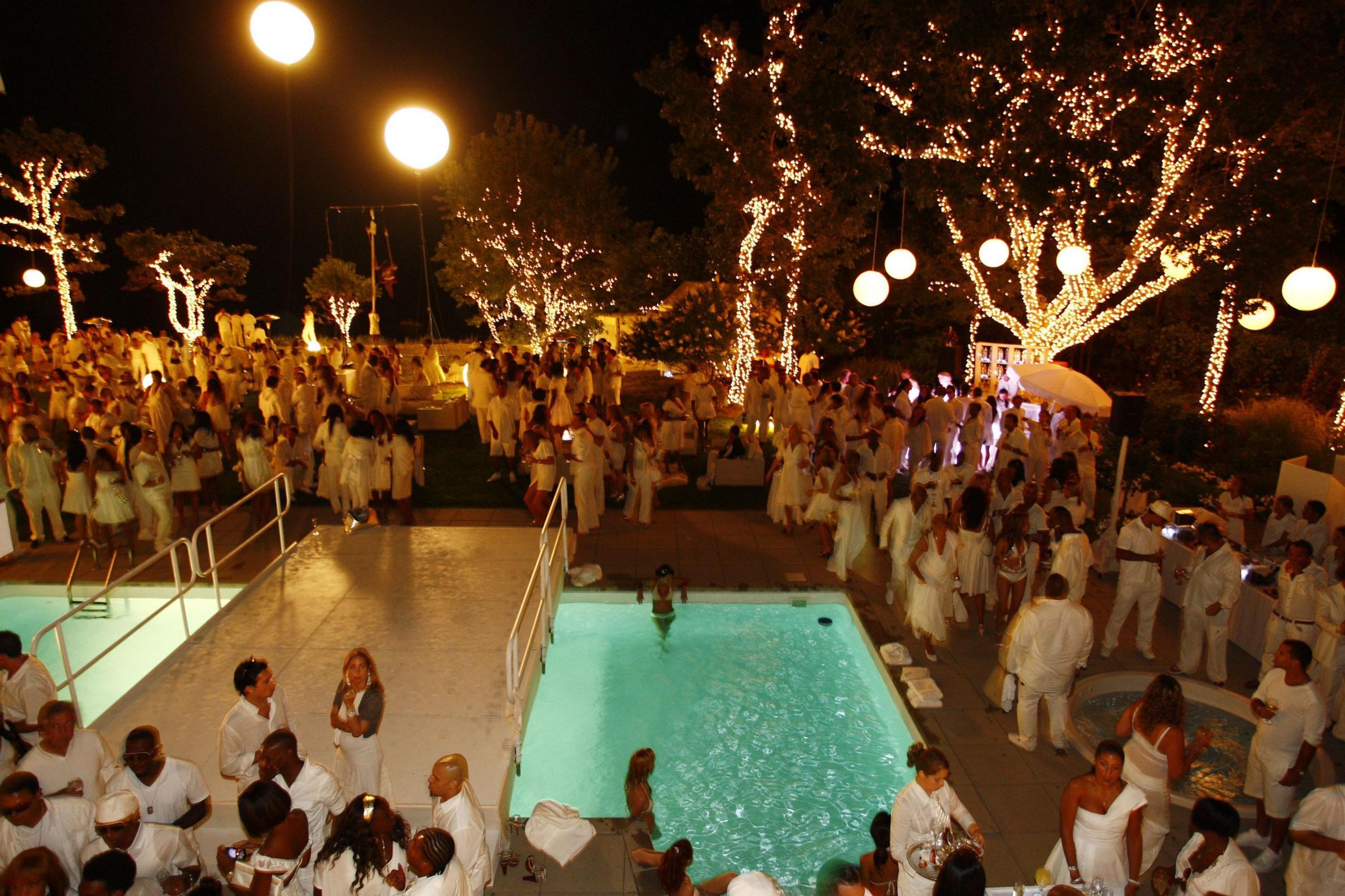 A White Party at the Hamptons in New York state, seen from a high vantage point, showing a large crowd all dressed in white, standing around a large swimming pool, which has a white bridge constructed over it. The tree trunks and branches are wrapped in fairy lights with large globe pendant lights hanging from some of them.