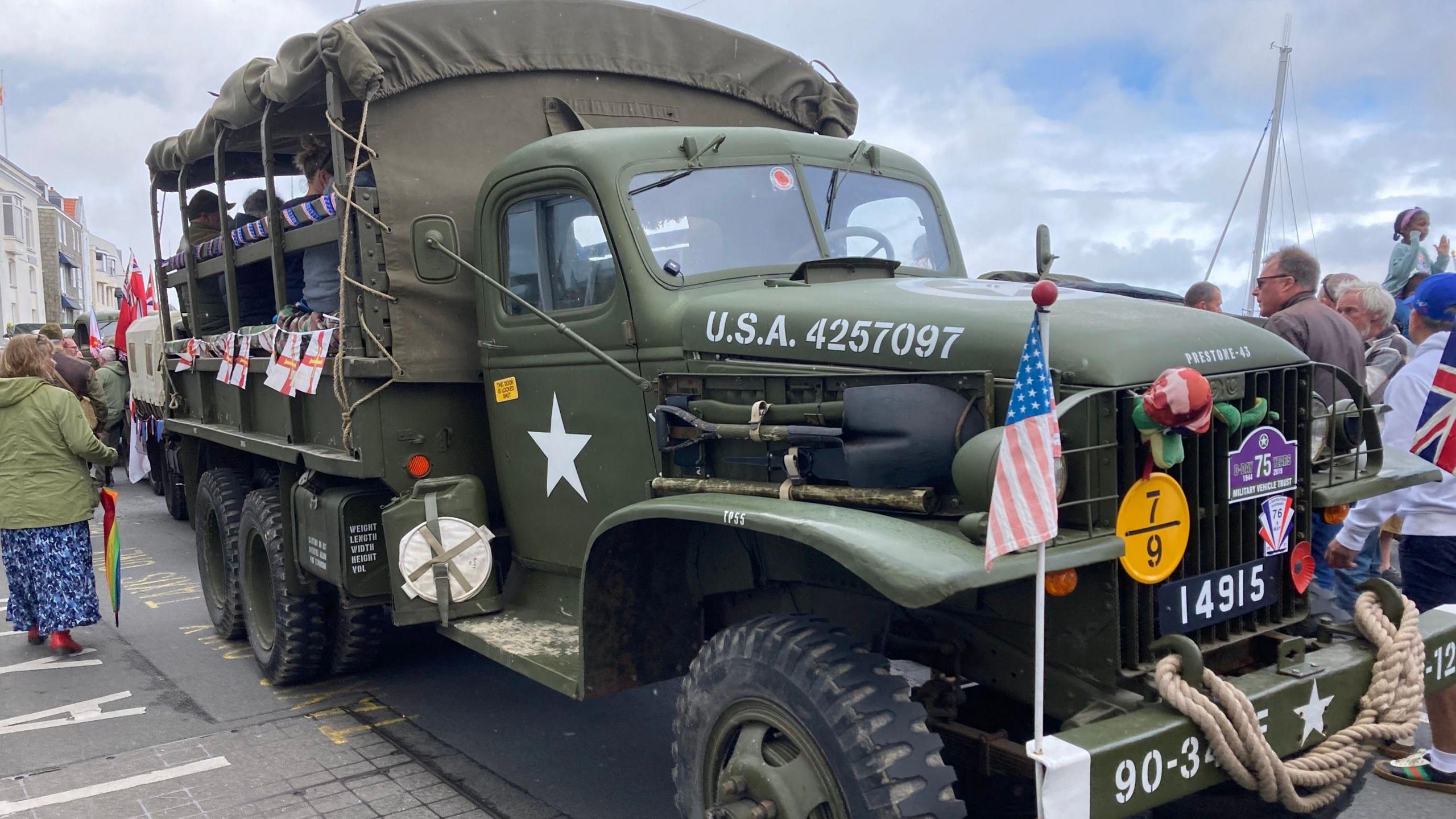 A USA military troop transport lorry. It has a canvas roof stretched over the seated area at the back. It is khaki green and has USA 4257097 printed in white on the large high bonnet and a five-pointed star in white on the passenger door. There is a USA flag tied to the bumper along with a Remembrance poppy and a length of looped rope. There are onlookers surrounding the lorry. 