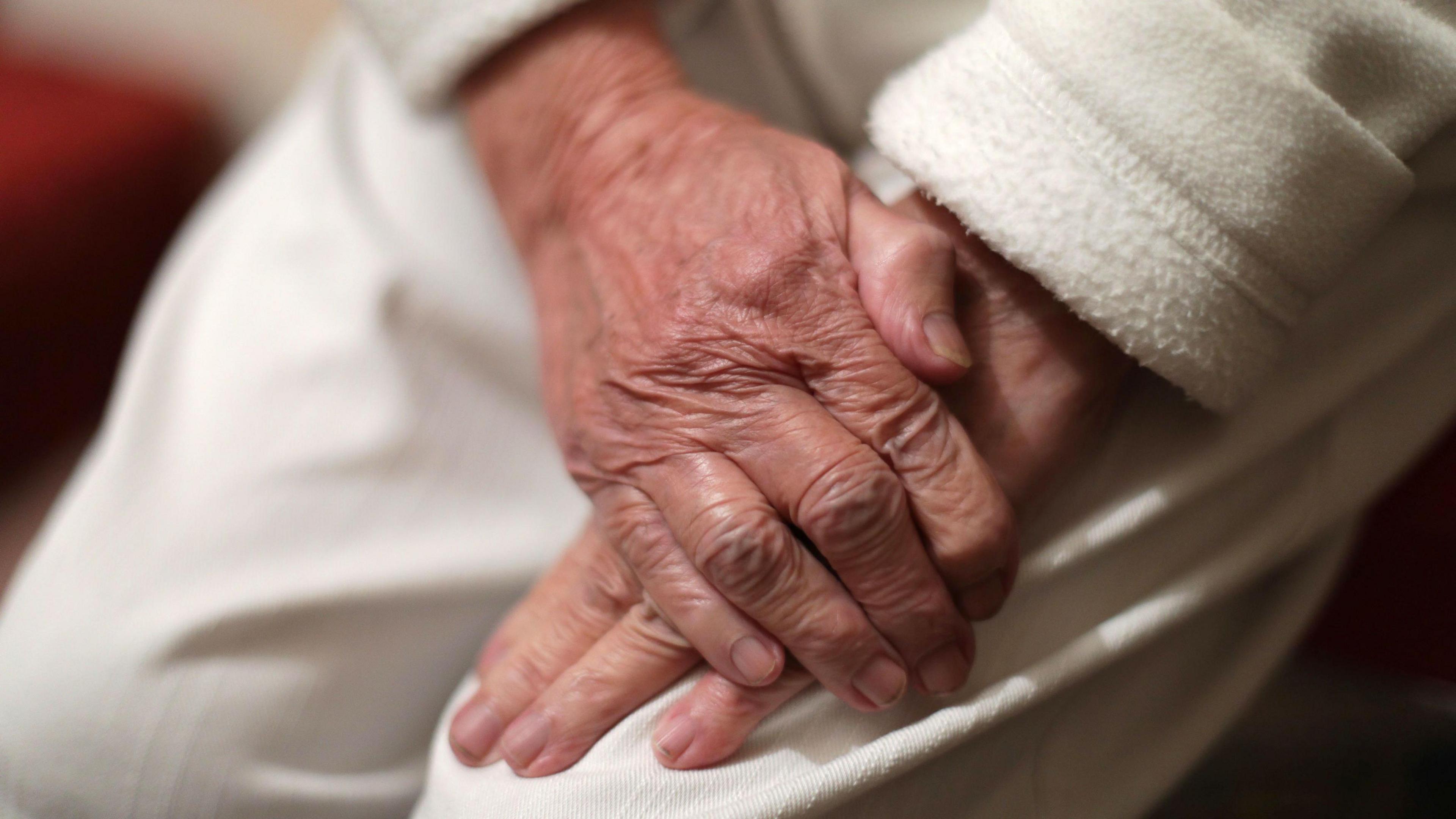 A close up of an elderly person's hands rest on white trousers. 