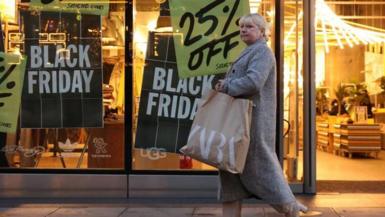 Woman carrying shopping bag walking past a shop covered in posters advertising Black Friday deals