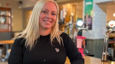 A young woman with long, blonde hair wearing a black pullover smiles as she sits at the bar of the pub she manages. The lights of the pumps on the bar twinkle behind her. 