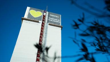 Grenfell Tower wrapped in white sheeting, with a large banner at the top displaying a green heart and the words "Grenfell forever in our hearts." The sky is clear and blue, and blurred dark leaves frame the right side of the image.