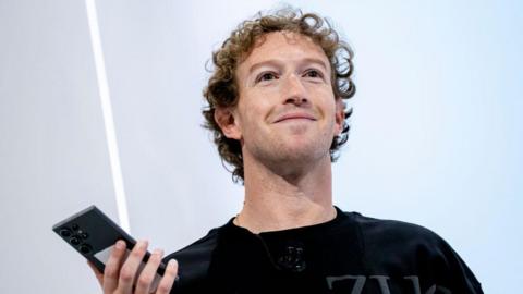 A smiling Mark Zuckerberg stands on stage holding up a smartphone