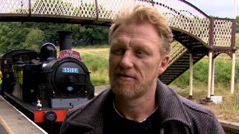 Kevin McKidd head and shoulders turned to the side talking to an interviewer. In the background there is a steam train and a bridge going over train tracks.