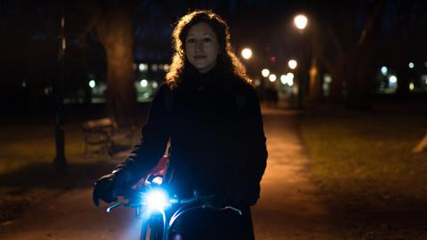 A woman holding her bike in the dark in a park. She wears a winter coat and gloves, and the bike has a bright bluish light, contrasting with the orangey lights in the park.