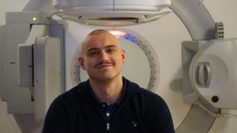 Max Vardy, a man with a bald head, pictured in front of a radiography machine. There is a green laser light running down his face.   