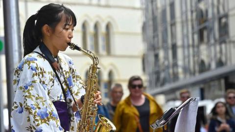 A girl wearing a white kimono with a pattern of blue flowers and green leaves plays a saxophone. In the background, people are walking along a city street.